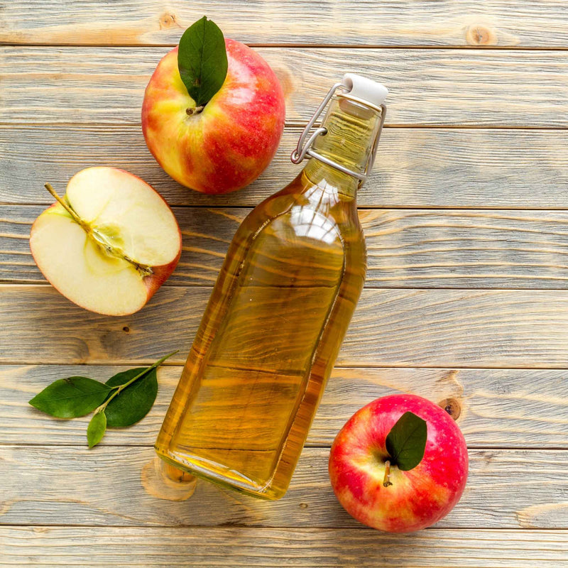 Bouteille de vinaigre de cidre non filtré avec la mère, sur une table en bois, entourée de pommes et d’herbes médicinales.